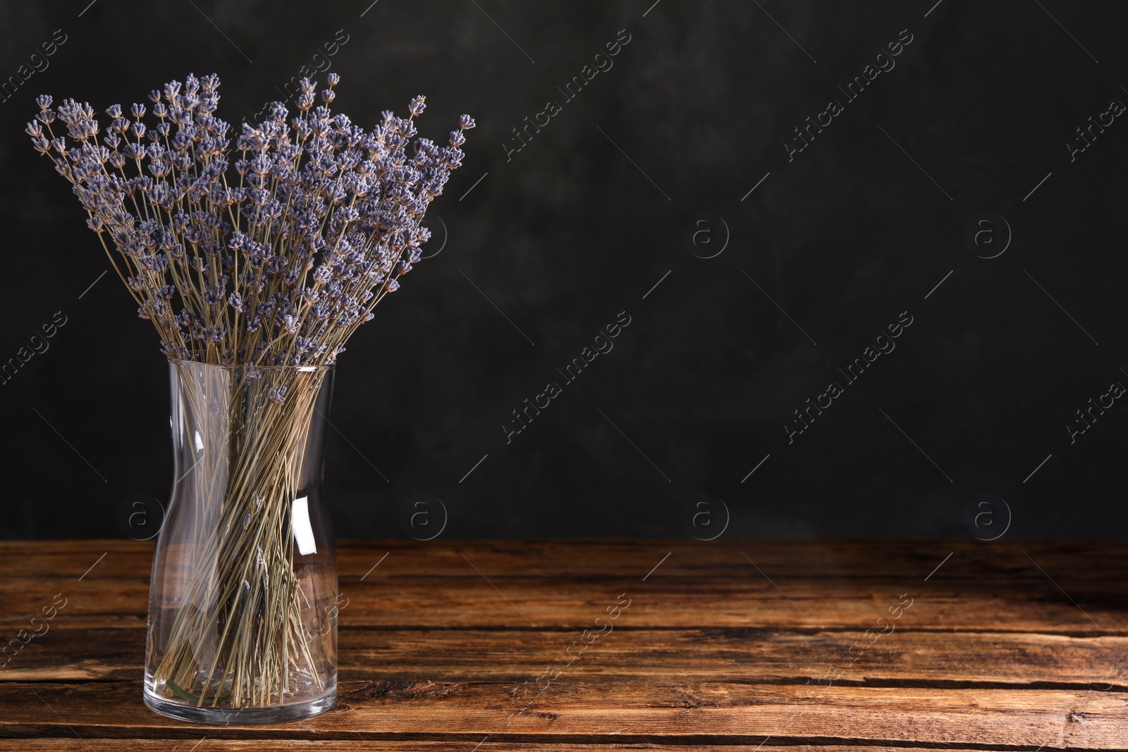 Photo of Dried lavender flowers in glass vase on wooden table against dark background. Space for text