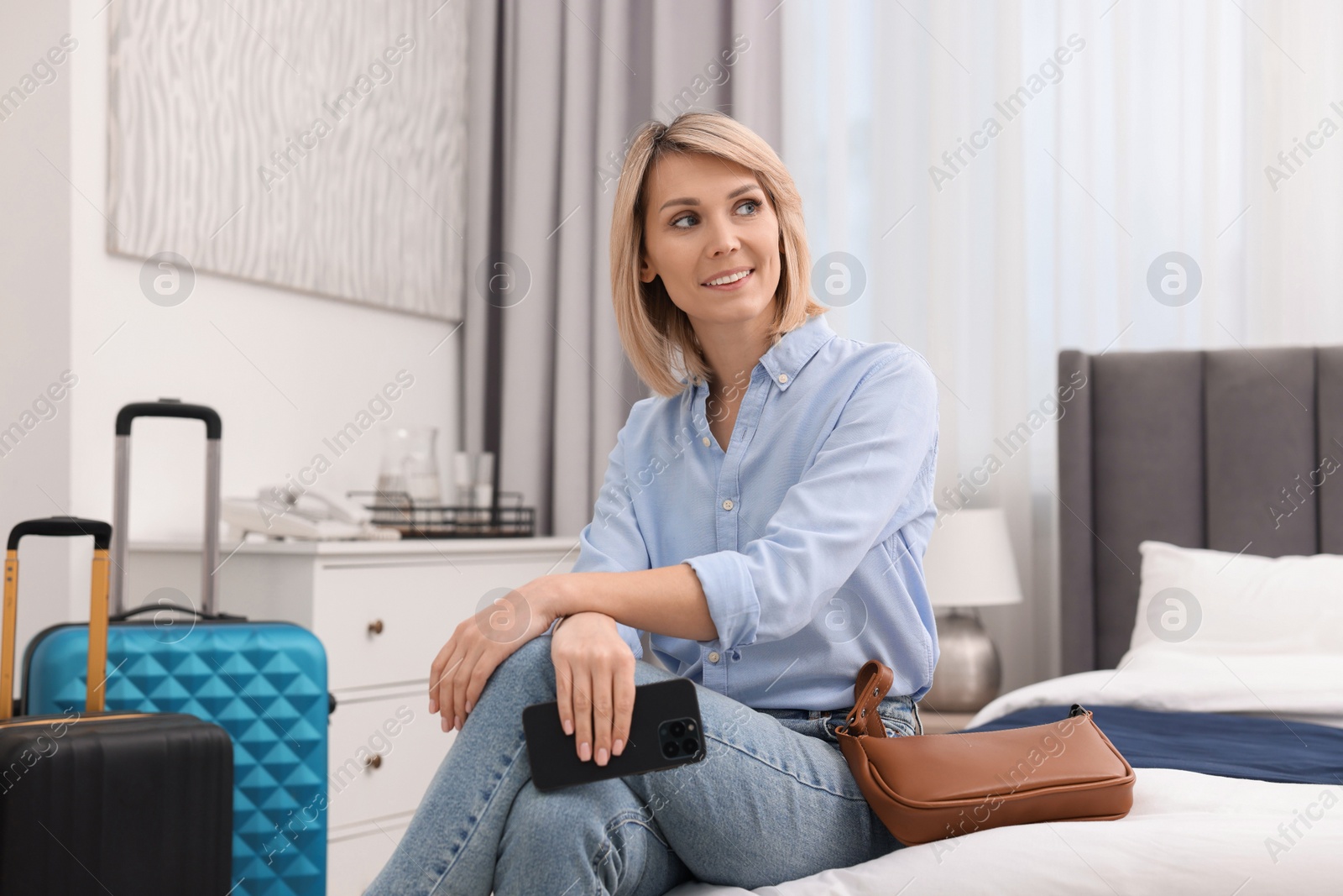 Photo of Smiling guest with smartphone on bed in stylish hotel room