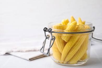 Photo of Tasty fresh yellow baby corns in glass jar on white tiled table, closeup. Space for text