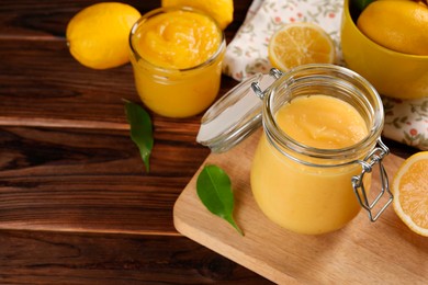 Photo of Delicious lemon curd in glass jars, fresh citrus fruits and green leaves on wooden table, space for text