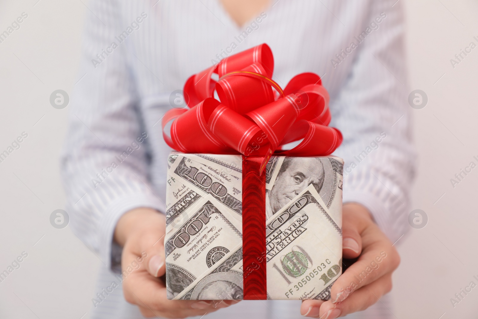 Photo of Woman holding gift box wrapped in dollars on light background, closeup