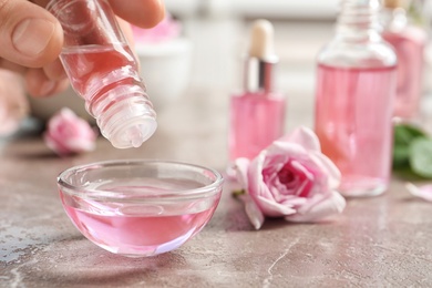 Woman dripping rose essential oil into bowl on table, closeup. Space for text