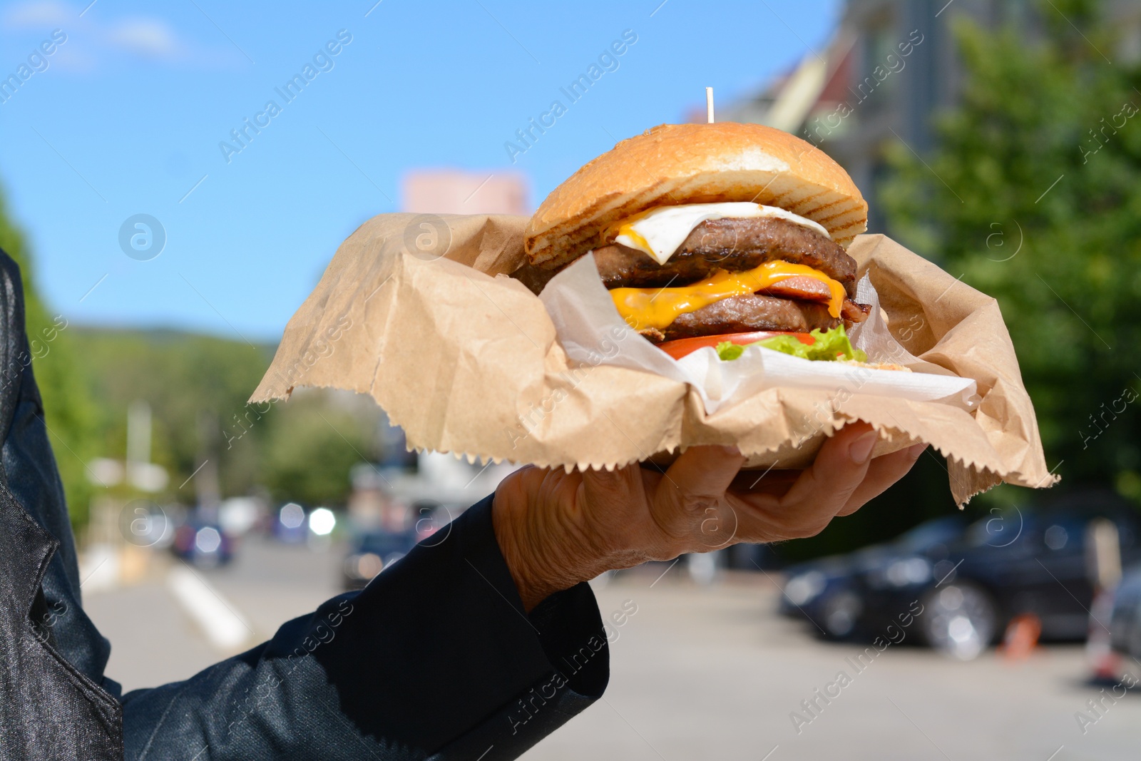 Photo of Woman holding delicious burger in paper wrap on city street, closeup