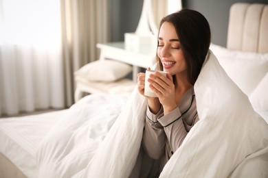 Photo of Beautiful young woman wrapped with soft blanket holding cup of hot drink on bed at home