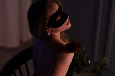 Photo of Elegant woman in black eye mask with rose indoors in evening