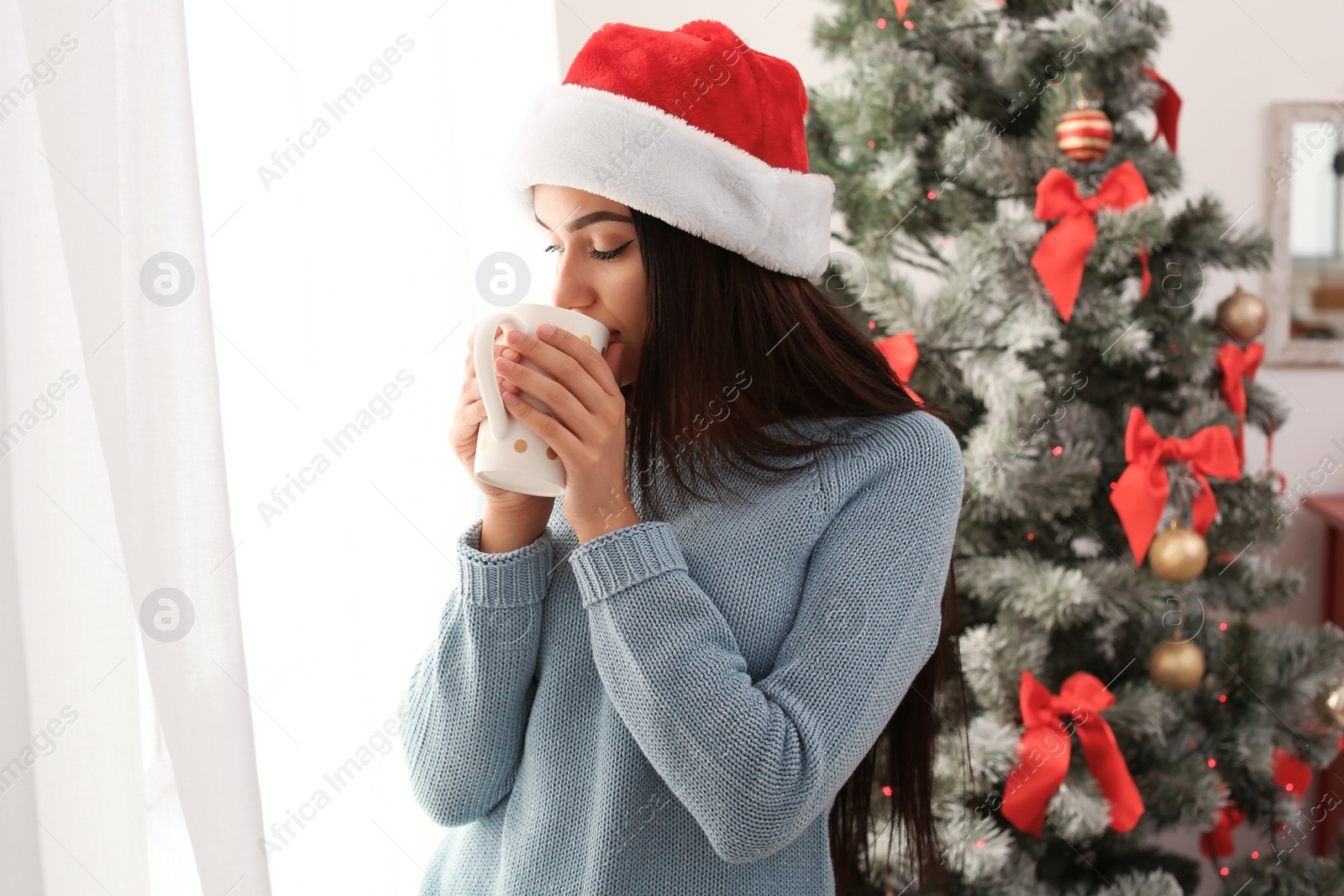 Photo of Beautiful young woman in Santa hat with cup of drink near Christmas tree at home