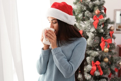 Beautiful young woman in Santa hat with cup of drink near Christmas tree at home