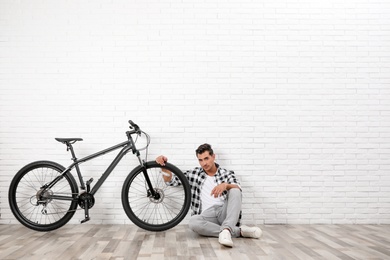 Handsome young man with modern bicycle near white brick wall indoors