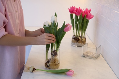 Photo of Woman putting tulips with bulbs into vase at table indoors, closeup