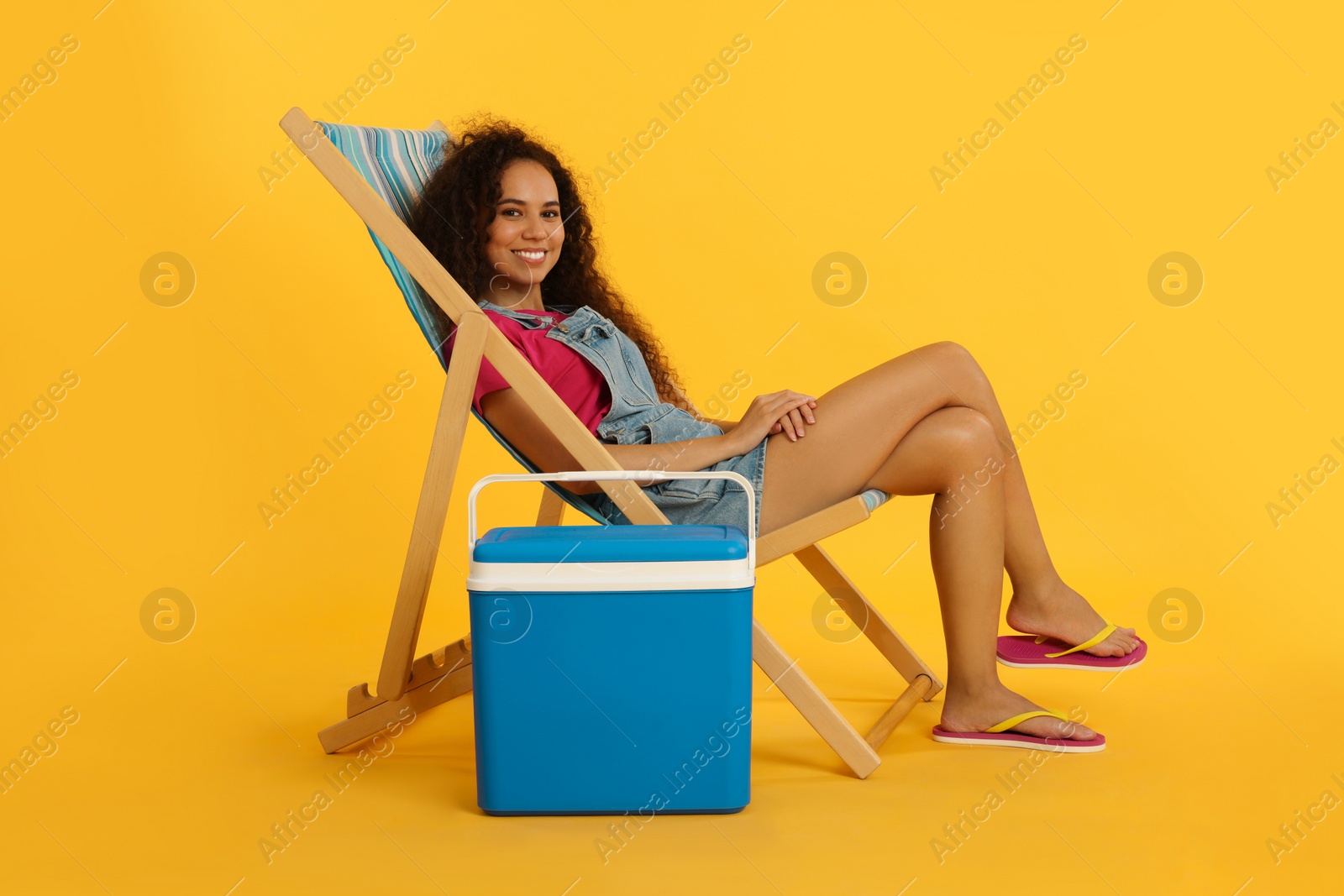 Photo of Happy young African American woman resting in deck chair near cool box on yellow background