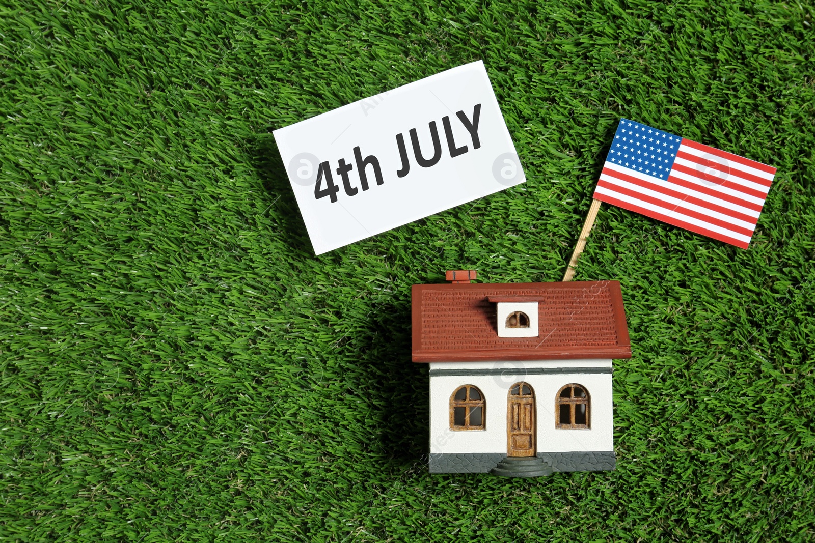 Photo of Flat lay composition with house model, USA flag and card on green grass, space for text. Happy Independence Day