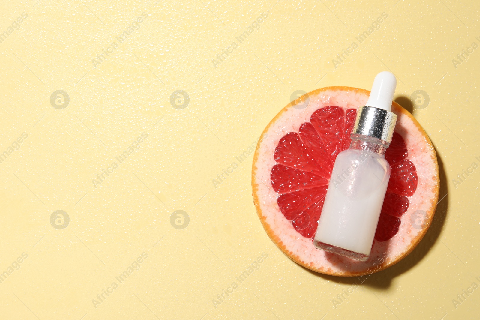 Photo of Bottle of cosmetic serum and grapefruit slice on wet yellow background, top view. Space for text