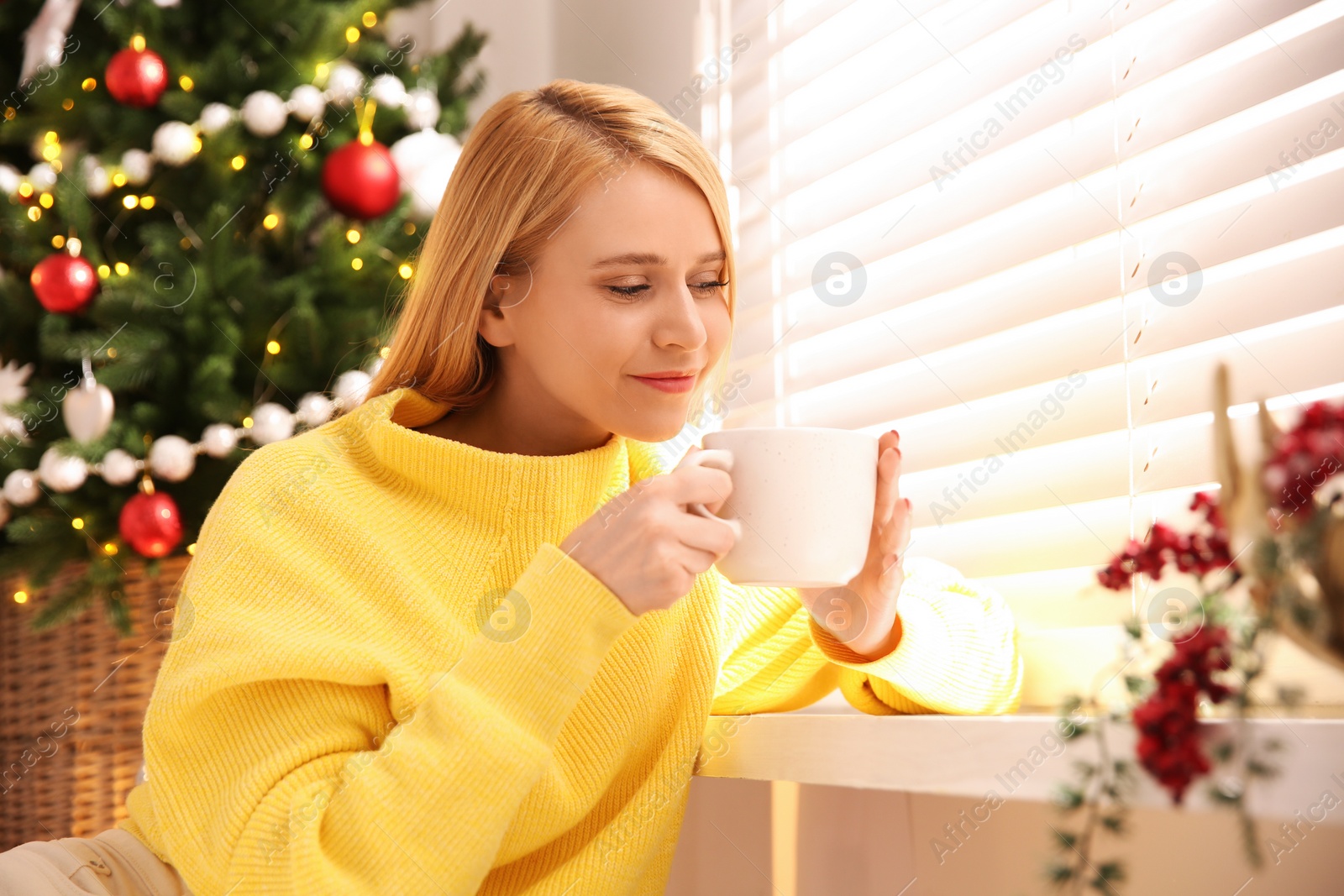 Photo of Young woman with cup of hot drink near Christmas tree at home