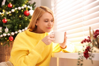 Photo of Young woman with cup of hot drink near Christmas tree at home