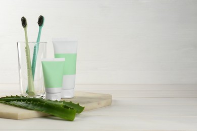 Photo of Tubes of toothpaste, toothbrushes and fresh aloe vera leaves on white wooden table. Space for text