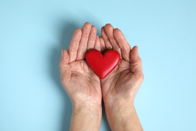 Elderly woman holding red heart in hands on light blue background, top view