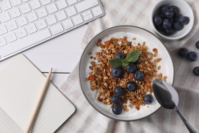 Photo of Delicious granola with blueberries in bowl, stationery and computer keyboard on white wooden table, flat lay