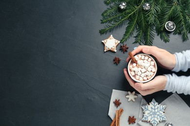 Photo of Woman holding cup of delicious hot chocolate with marshmallows and cinnamon at black table, top view. Space for text