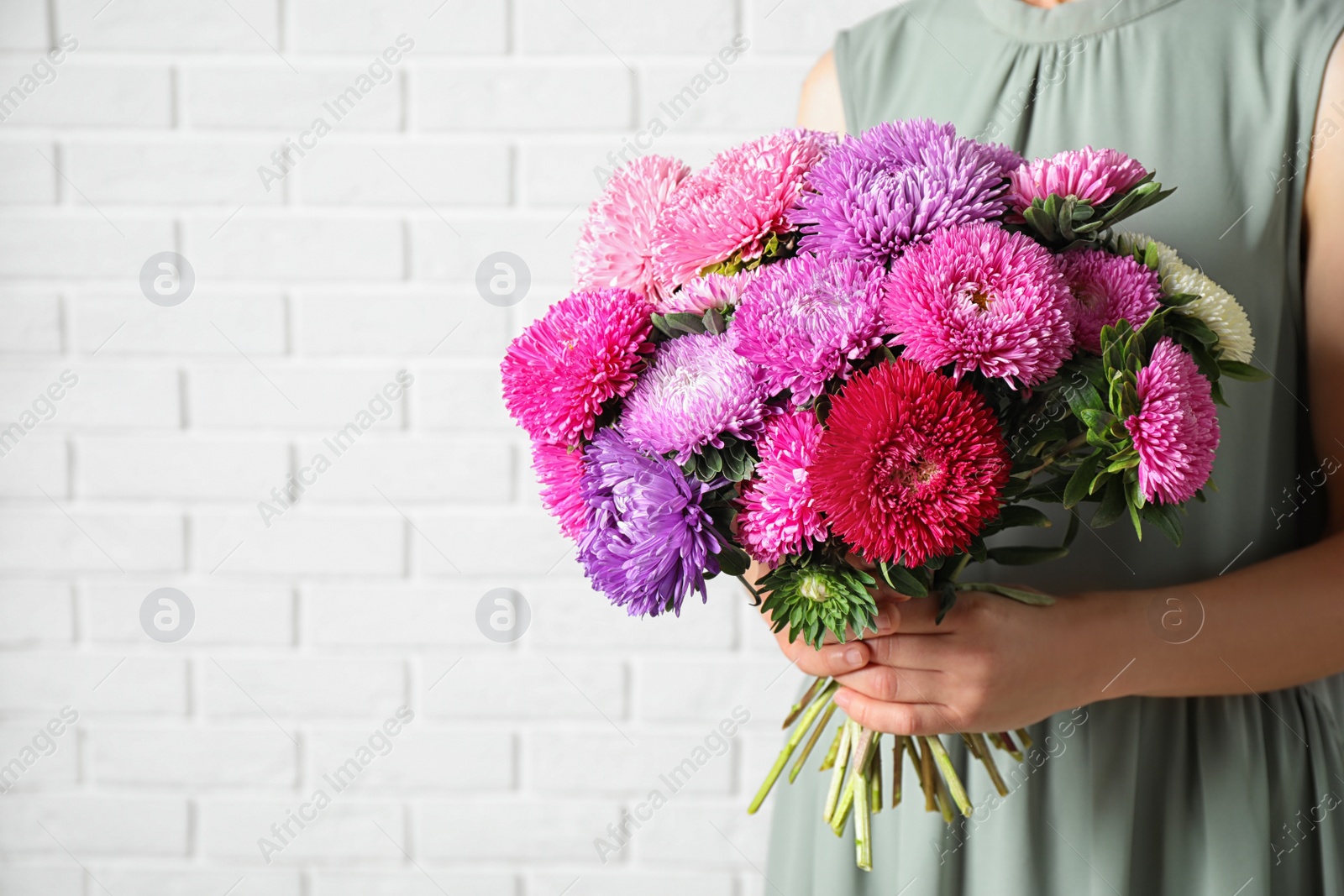 Photo of Woman holding bouquet of beautiful aster flowers against white brick wall, closeup. Space for text