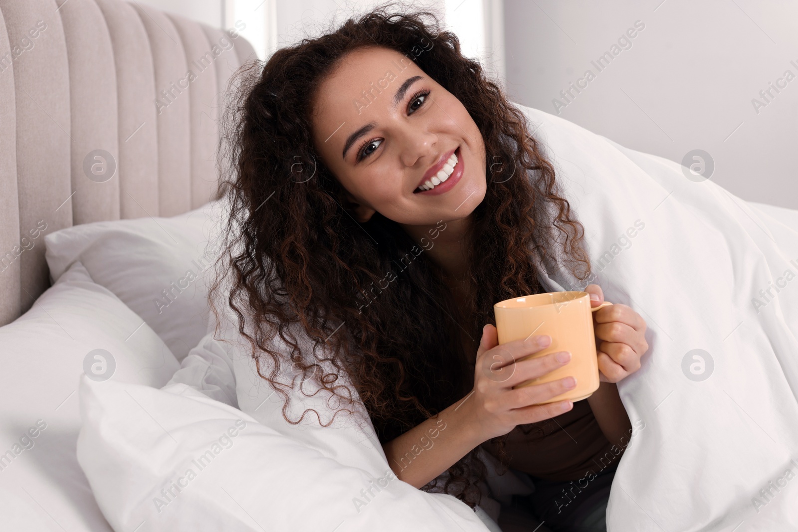 Photo of Happy African American woman with cup of drink in bed at home