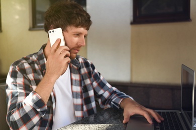 Handsome man talking on phone while using laptop at table in cafe