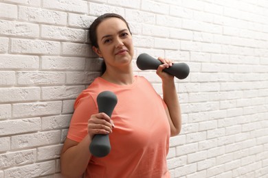 Happy overweight woman doing exercise with dumbbells near white brick wall
