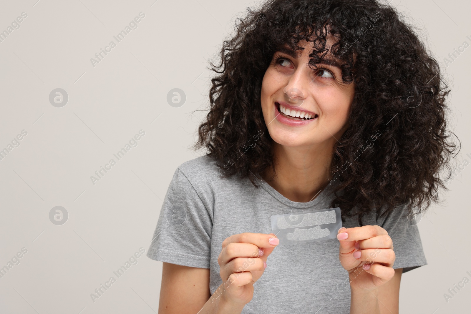 Photo of Young woman holding teeth whitening strips on light grey background, space for text