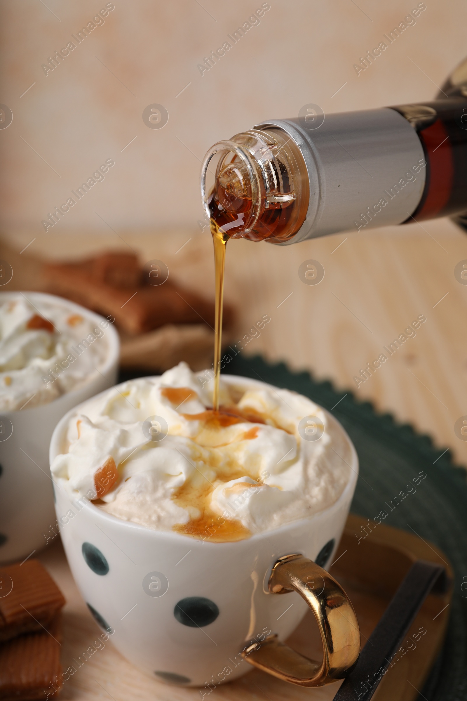 Photo of Pouring delicious caramel syrup into cup with coffee and whipped cream at wooden table, closeup