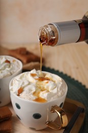 Pouring delicious caramel syrup into cup with coffee and whipped cream at wooden table, closeup