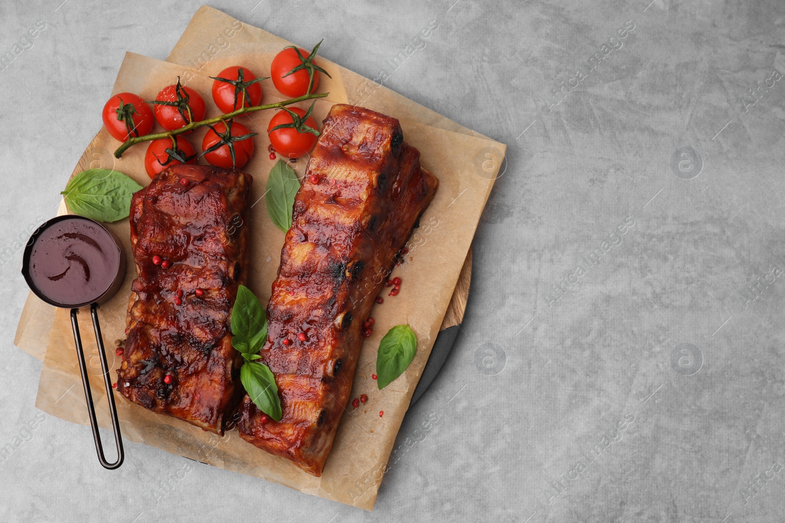 Photo of Tasty roasted pork ribs served with sauce, basil and tomatoes on grey table, top view. Space for text