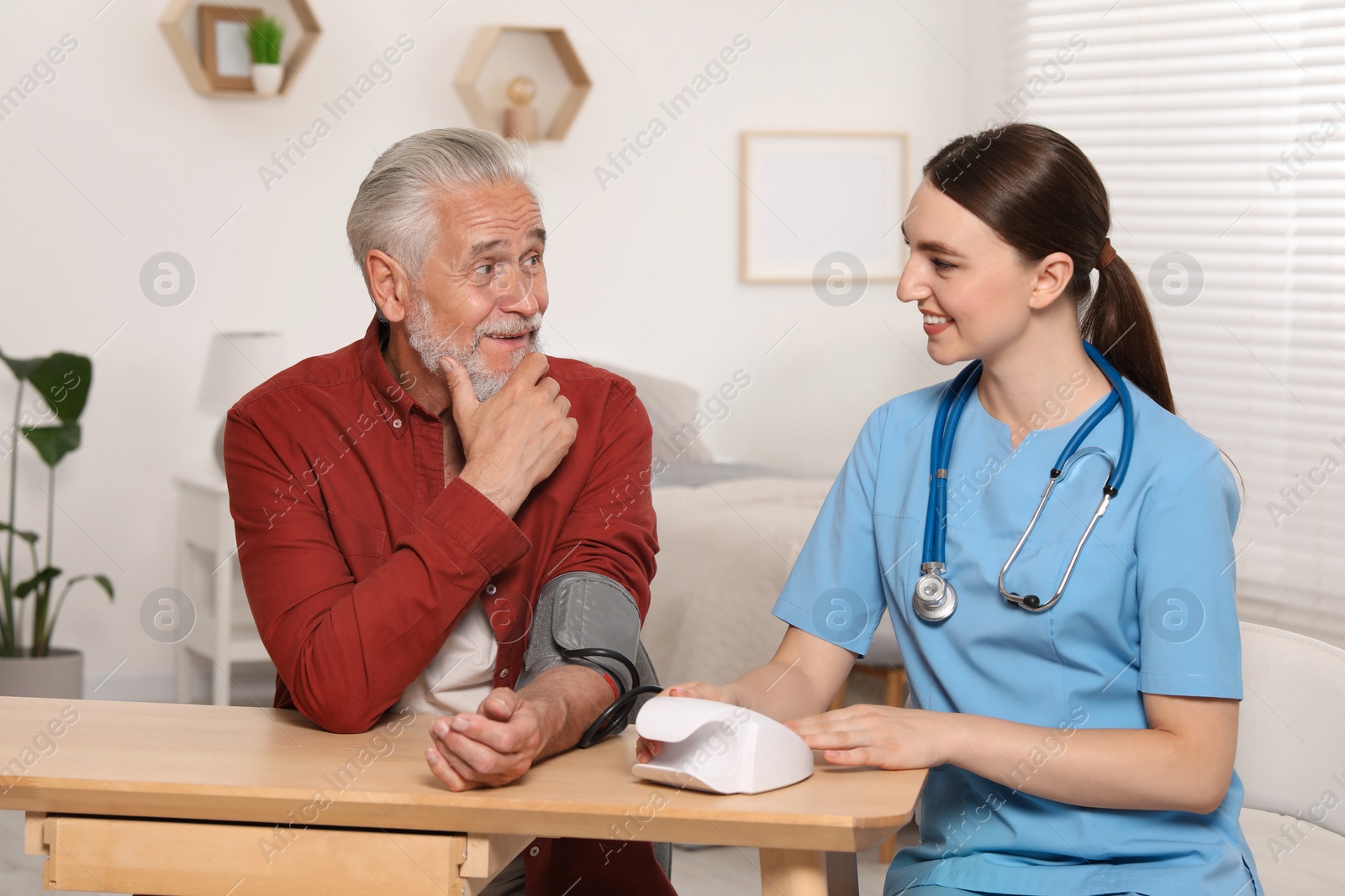 Photo of Young healthcare worker measuring senior man's blood pressure at wooden table indoors