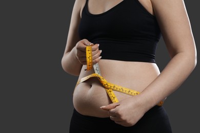 Woman measuring belly with tape on grey background, closeup. Overweight problem