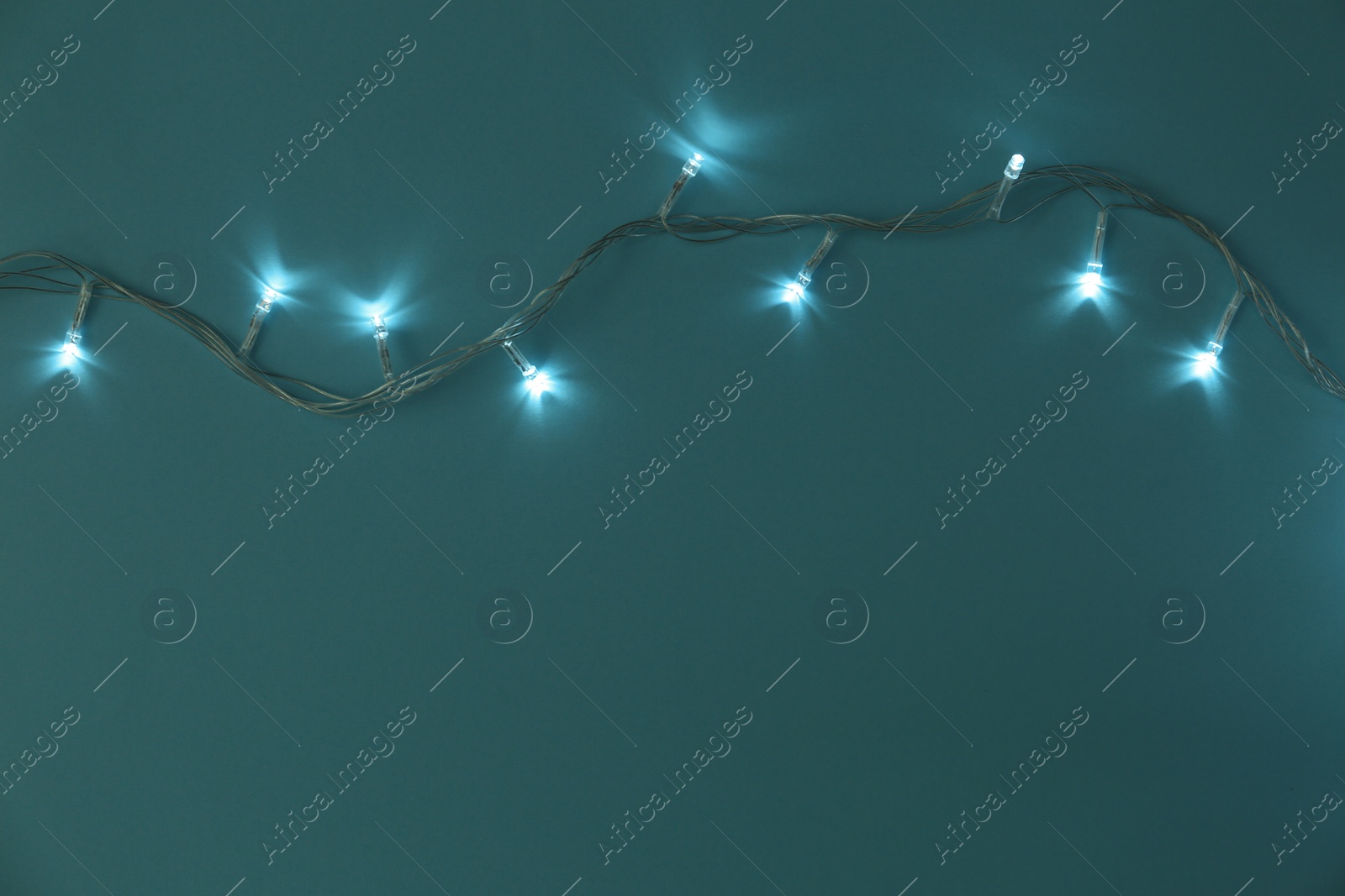 Photo of Glowing Christmas lights on turquoise background, top view. Space for text