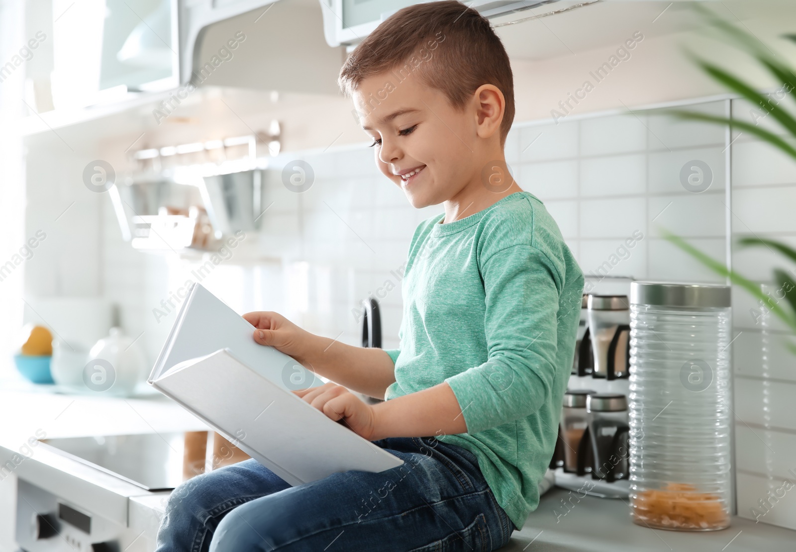 Photo of Cute little boy reading book in kitchen at home