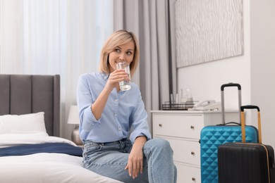 Smiling guest drinking glass of water on bed in stylish hotel room