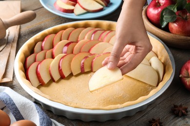 Woman putting apple slices into dish with raw dough at wooden table, closeup. Baking pie