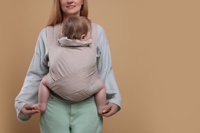Mother holding her child in sling (baby carrier) on light brown background, closeup. Space for text
