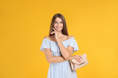Young woman wearing stylish dress with elegant clutch on yellow background