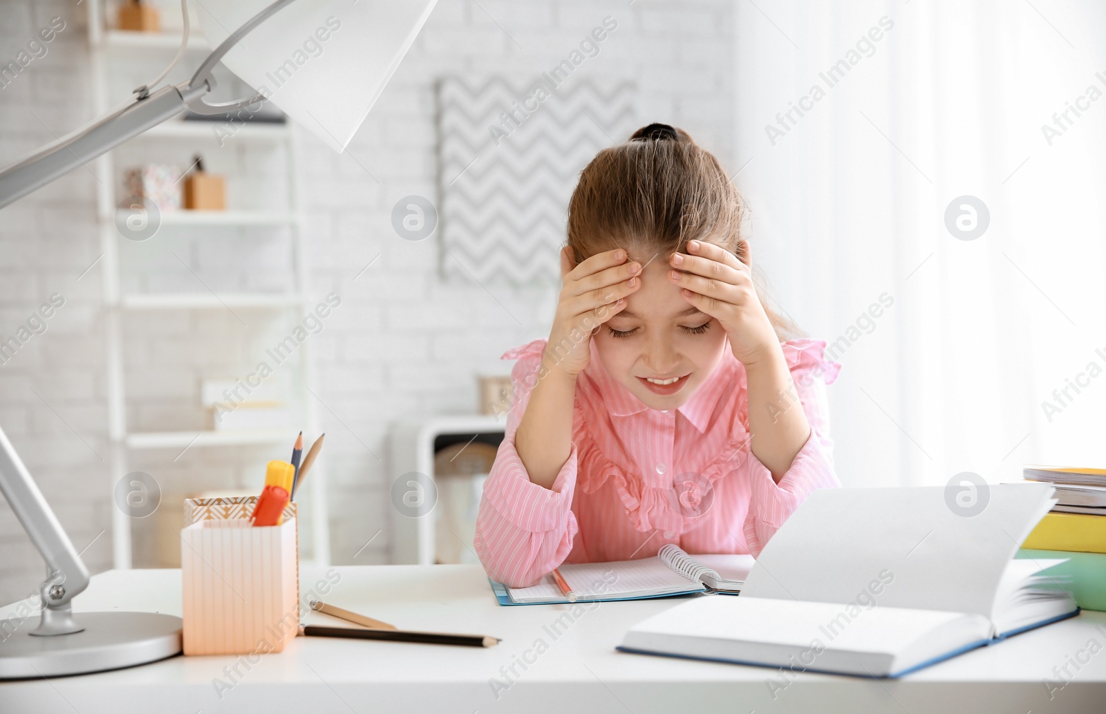 Photo of Little girl suffering from headache while doing homework at table indoors