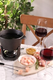 Photo of Fondue pot with oil, forks, raw meat pieces, glasses of red wine and other products on white marble table
