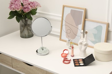 Mirror, cosmetic products, perfumes and vase with pink roses on white dressing table in makeup room