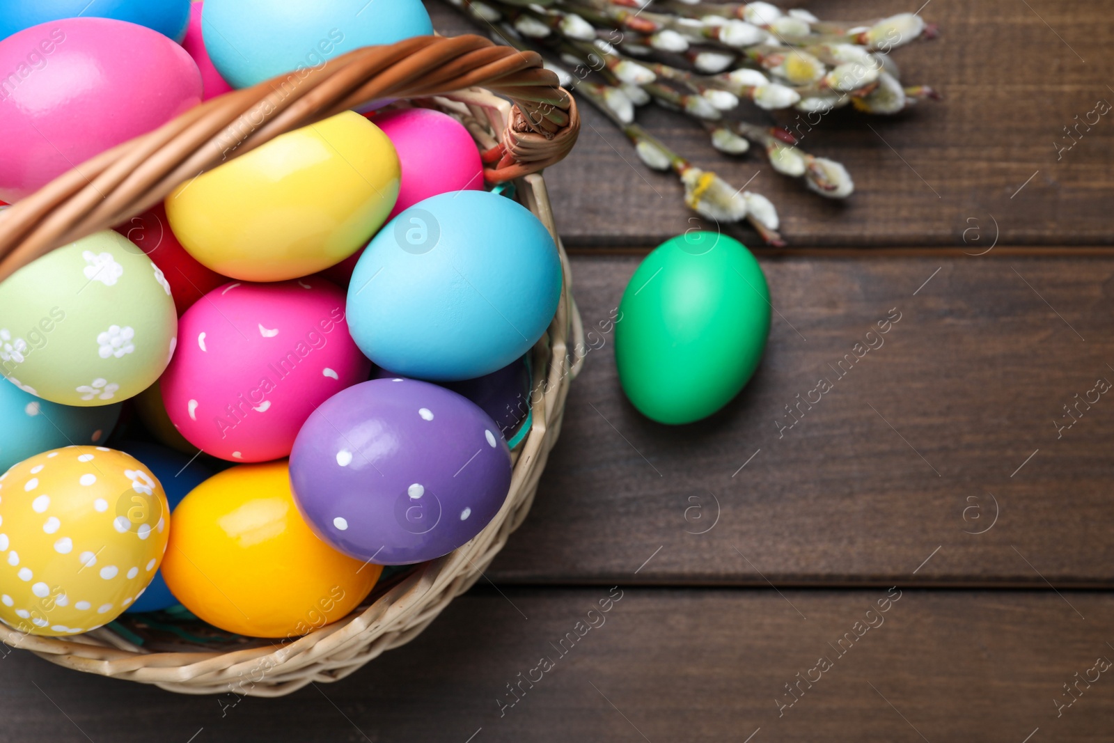 Photo of Colorful Easter eggs in wicker basket and willow branches on wooden table, flat lay. Space for text