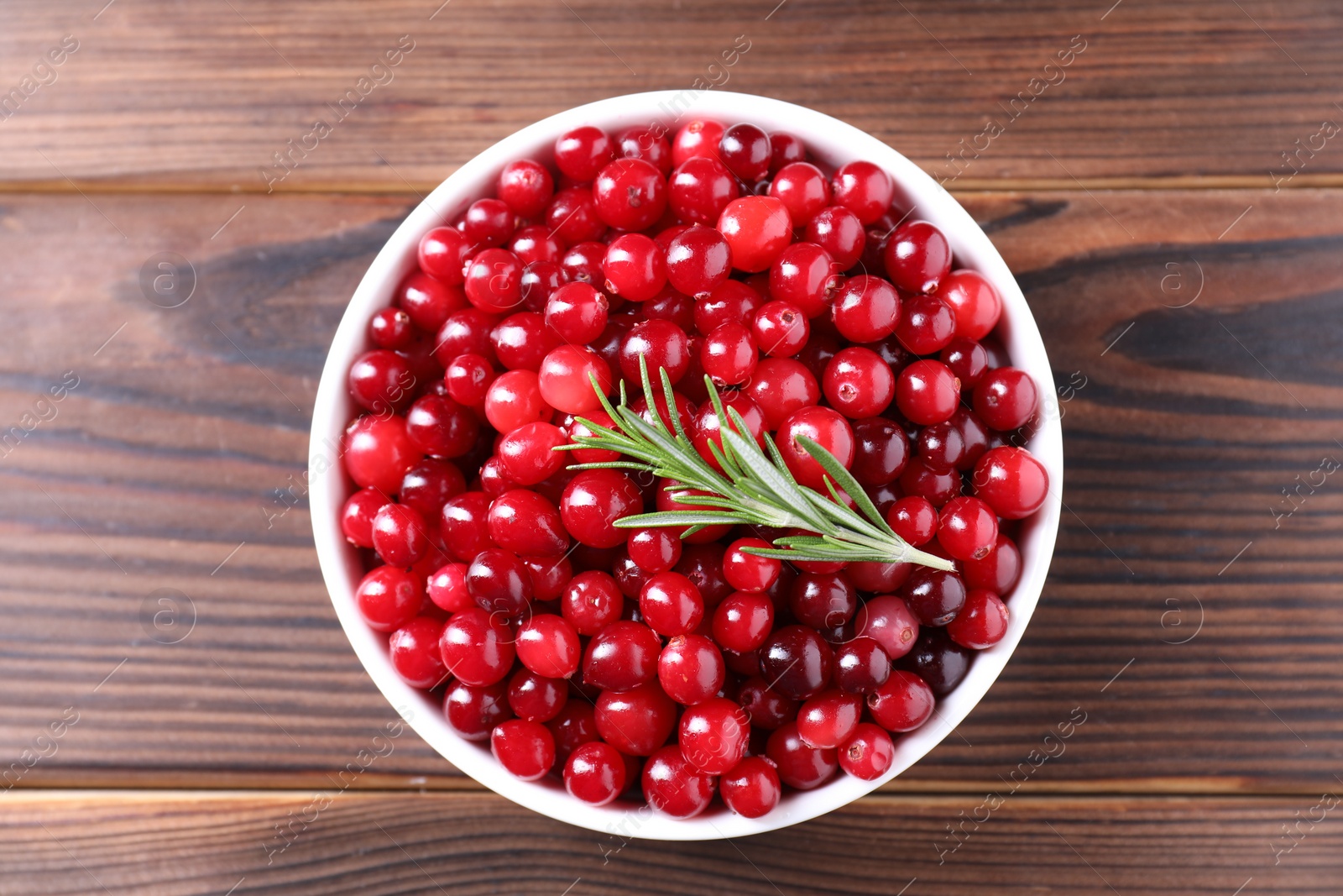 Photo of Fresh ripe cranberries and rosemary in bowl on wooden table, top view