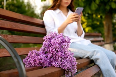 Photo of Young woman using mobile phone in park, focus on bunch of lilac flowers