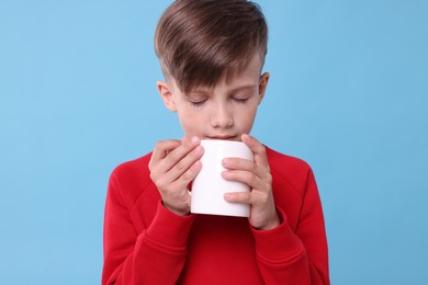 Photo of Cute boy drinking beverage from white ceramic mug on light blue background