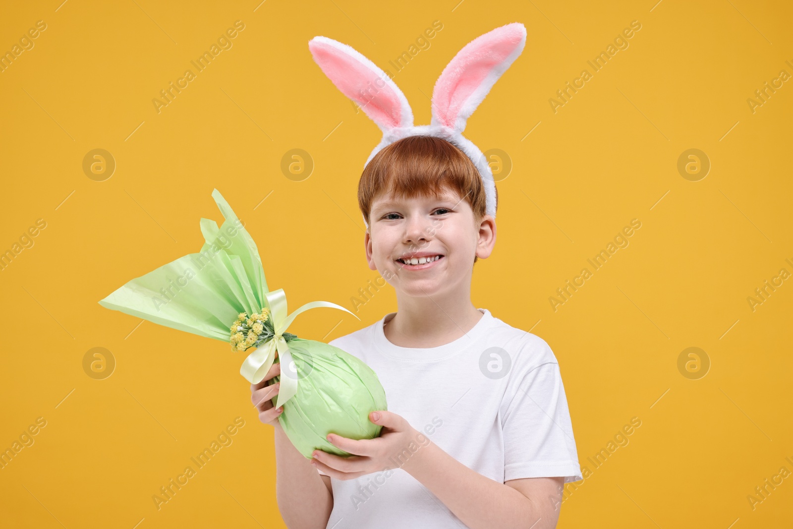 Photo of Easter celebration. Cute little boy with bunny ears and wrapped egg on orange background
