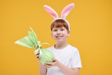 Photo of Easter celebration. Cute little boy with bunny ears and wrapped egg on orange background