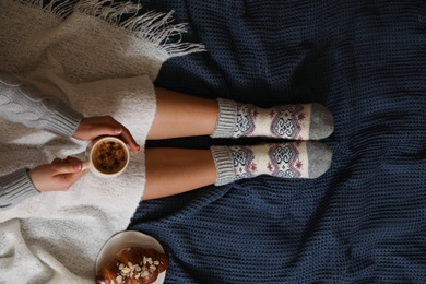 Photo of Woman in warm socks with cup of hot drink relaxing on knitted blanket, top view