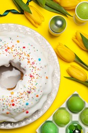 Easter cake with sprinkles, painted eggs and tulips on yellow background, flat lay