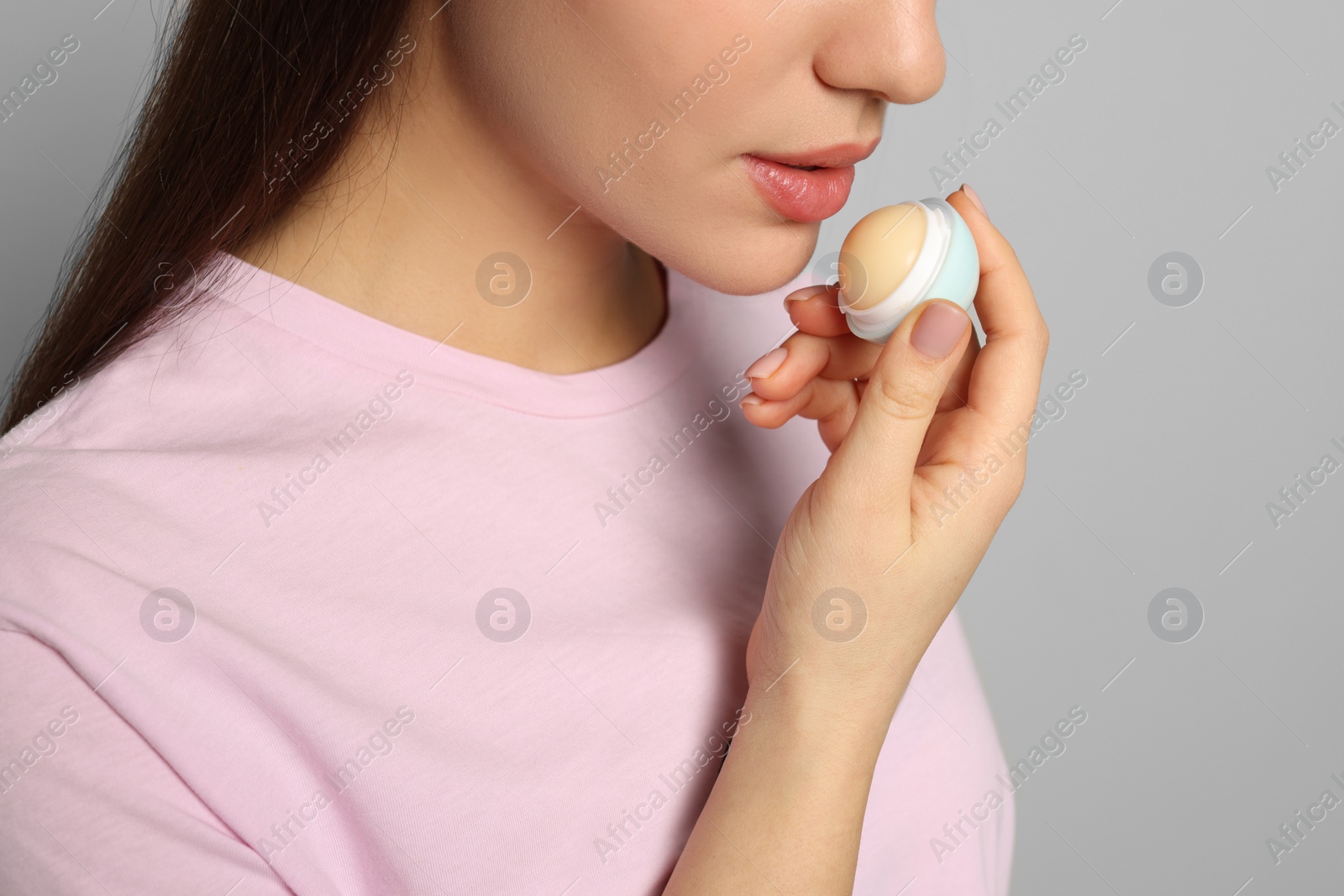 Photo of Young woman applying lip balm on grey background, closeup